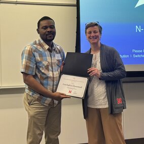 Two people standing indoors holding a certificate in a classroom setting.